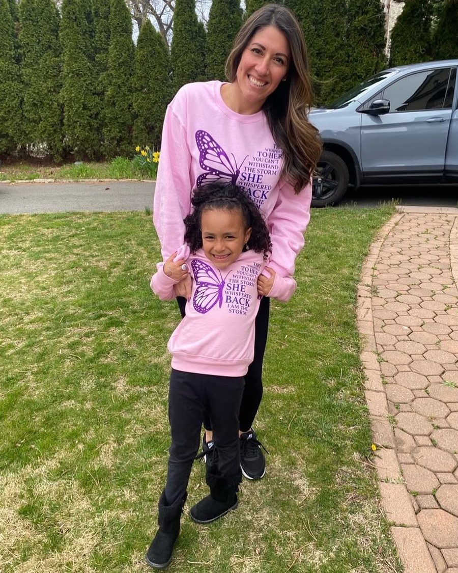 Deej smiling outdoors with a little girl, both in matching pink butterfly sweatshirts that read “She whispered back, I am the storm”